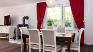 A dining area with a wooden table and white upholstered chairs, a fireplace, and red curtains. - Ferienhaus Yvonne in Wendorf im Bernstein Resort (Wendorf)