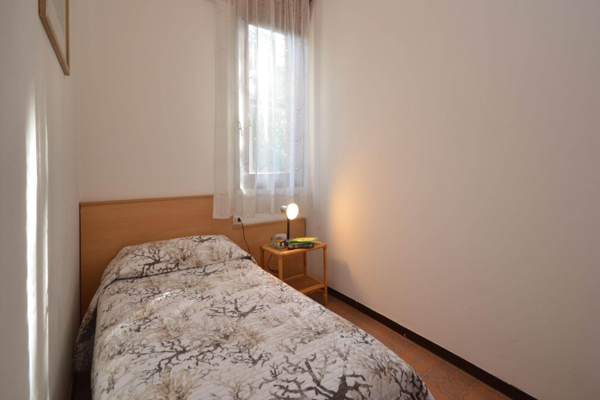 A bedroom with a bed, a bedside table with a lamp, and a window with sheer curtains.