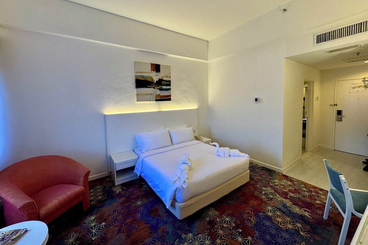 Une chambre d’hôtel avec un lit, un fauteuil rouge, une petite table et un tapis à motifs.