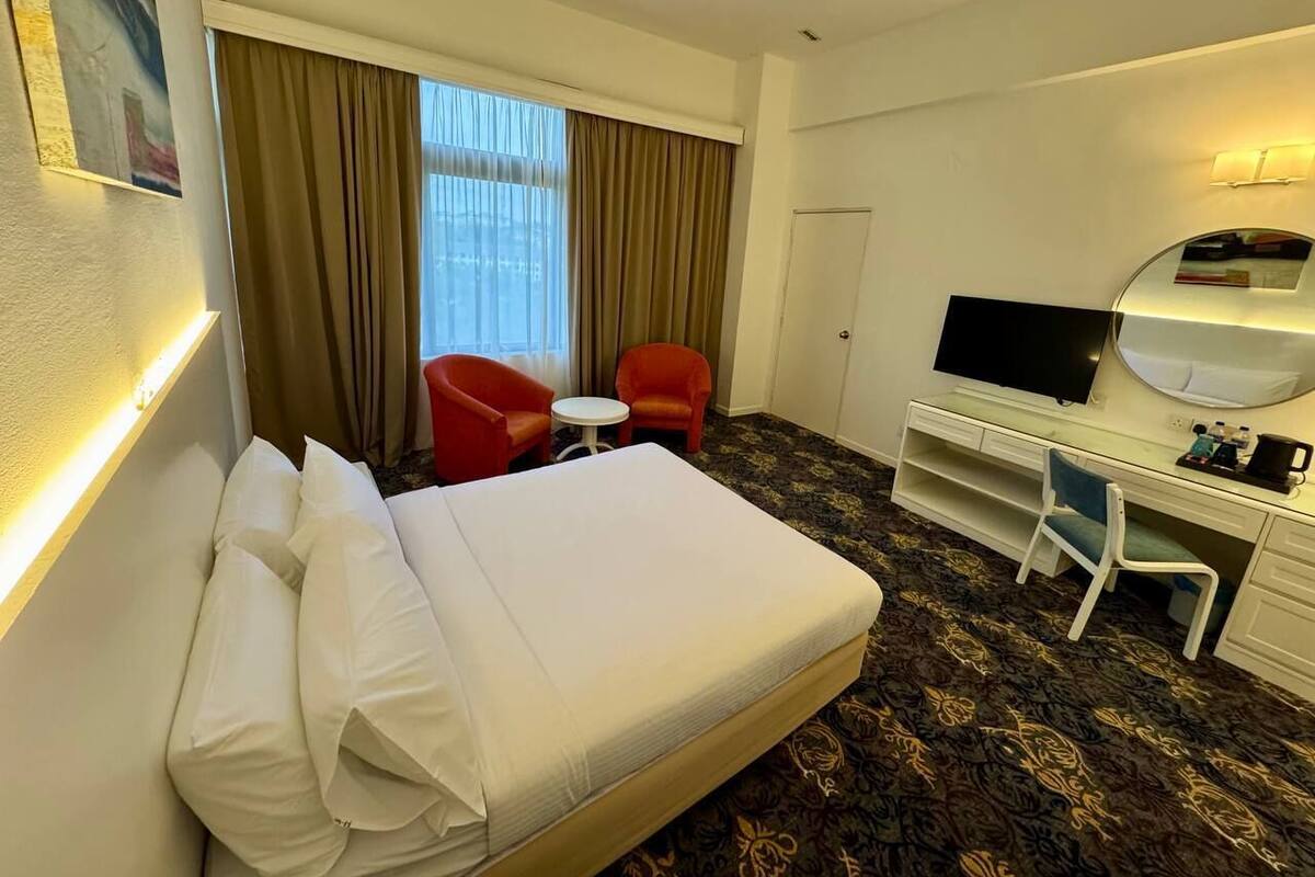 Une chambre d’hôtel avec un lit, deux fauteuils rouges, une petite table, un bureau avec une chaise, une télévision et une grande fenêtre avec des rideaux.