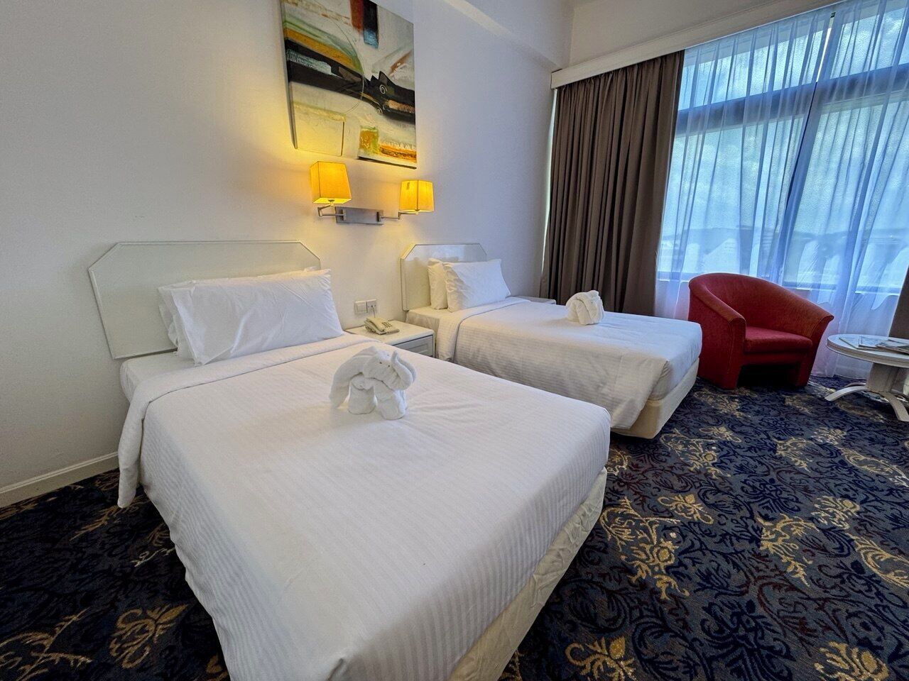 A hotel room with two beds, a red chair, a small table, and a painting on the wall.
