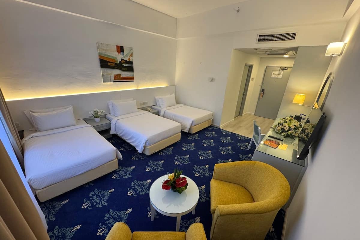 Une chambre d’hôtel avec deux lits, une petite table sur laquelle est posé un vase de fleurs, un fauteuil jaune et un bureau avec une lampe et des fleurs.