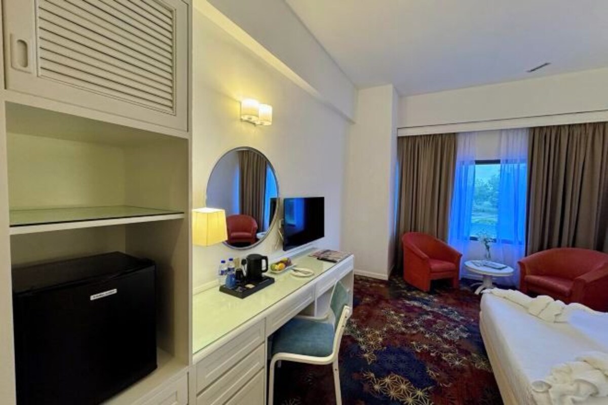 A hotel room with a bed, a desk with a TV, a chair, a small table, and a window with curtains.