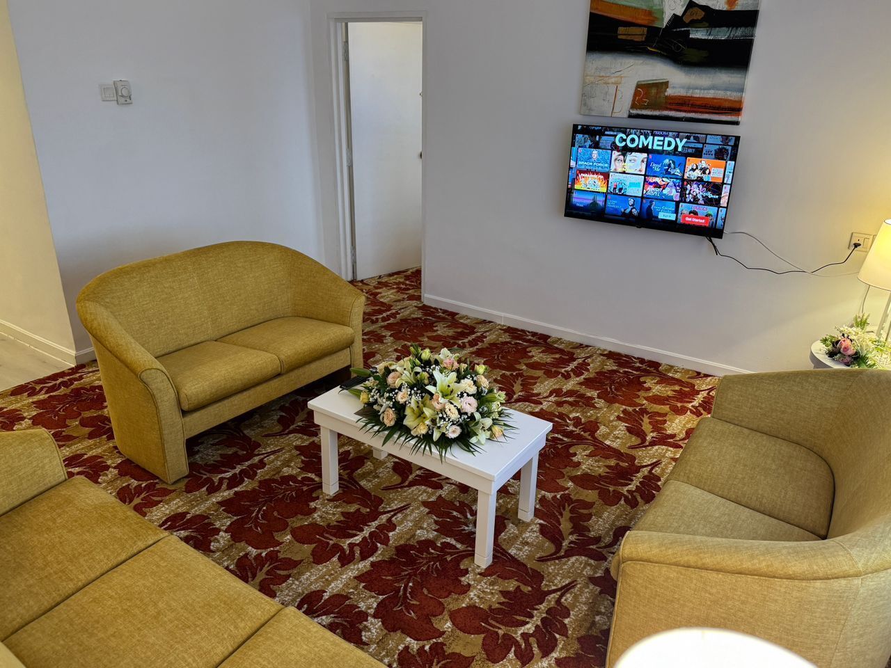 A living room with two yellow armchairs, a white coffee table with a floral arrangement, a red patterned carpet, and a TV mounted on the wall displaying a comedy channel.
