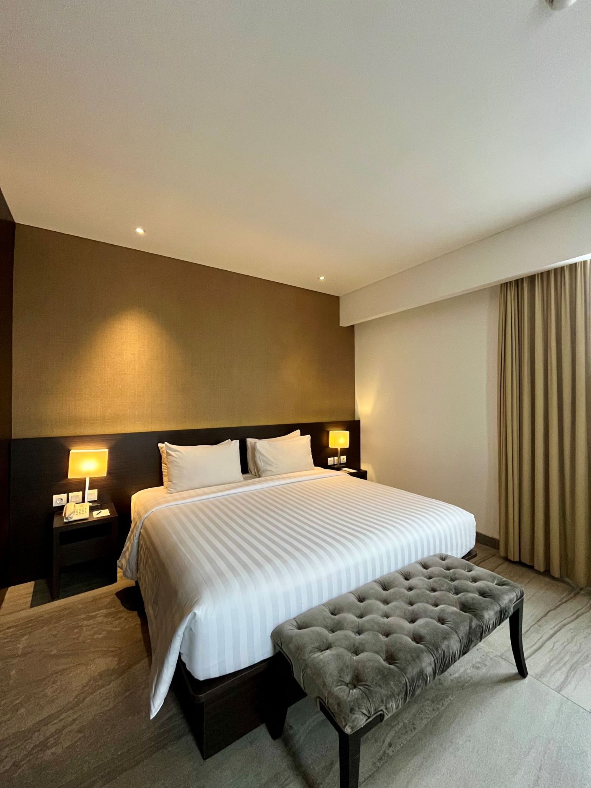 A hotel room with a large bed, a bedside table with a lamp, a bench, and a headboard.