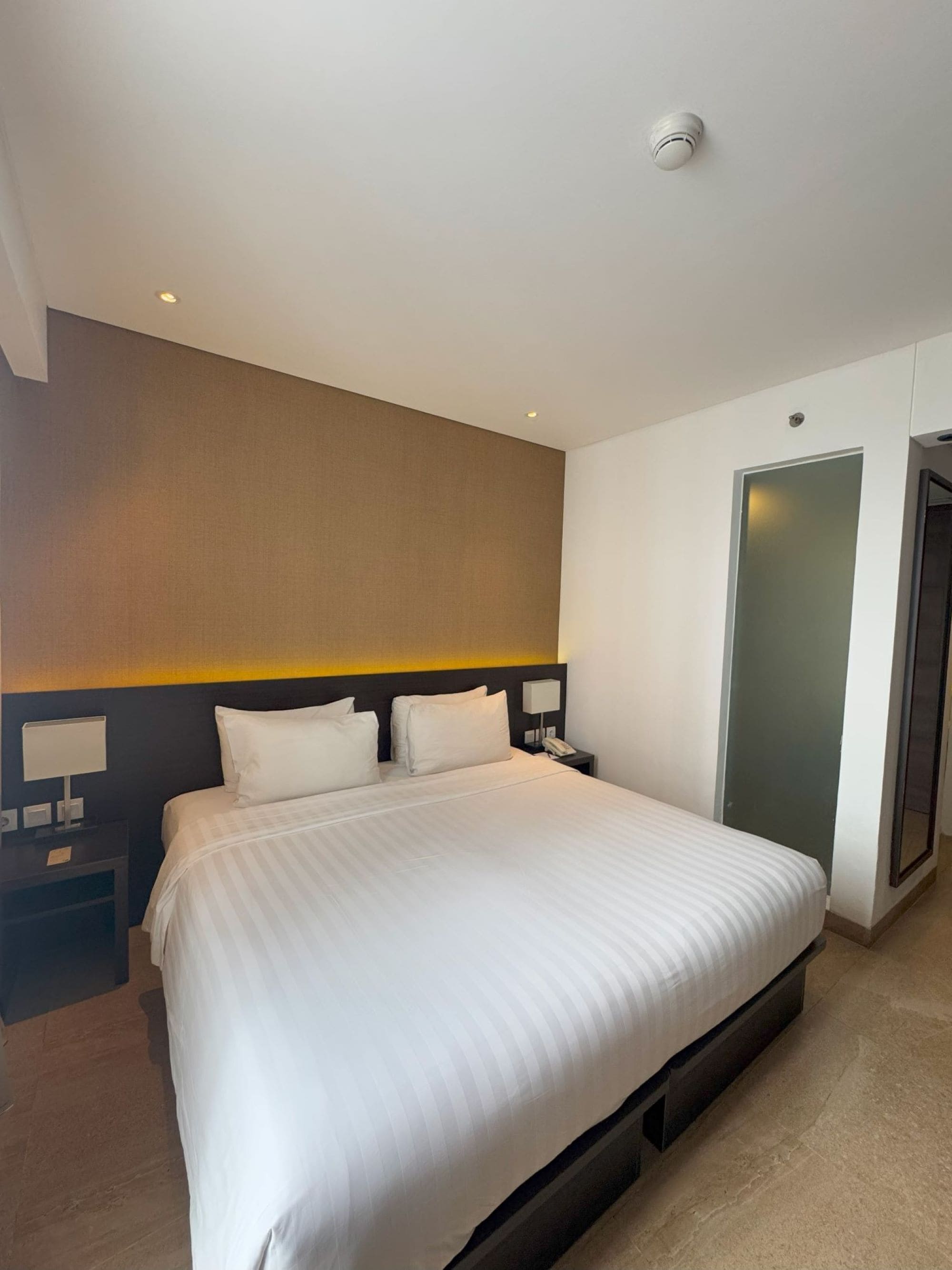 Quarto de hotel com uma cama grande, uma mesa de cabeceira com abajur, uma luz na parede e uma porta.