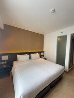 Quarto de hotel com uma cama grande, uma mesa de cabeceira com abajur, uma luz na parede e uma porta.