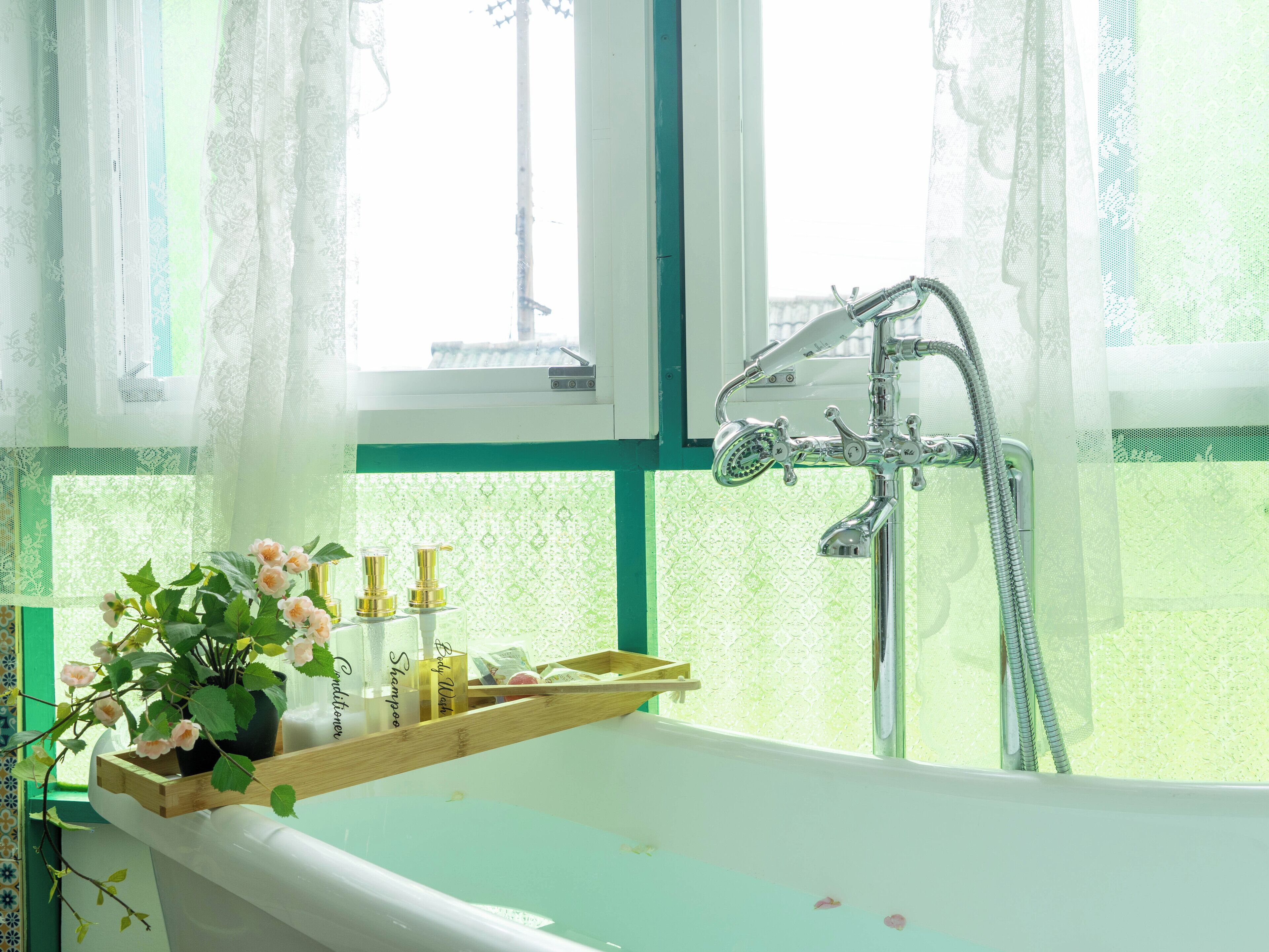 Ein Badezimmer mit einer Badewanne, einem Fenster mit Spitzenvorhängen und Pflegeprodukten auf einer Holztablett.