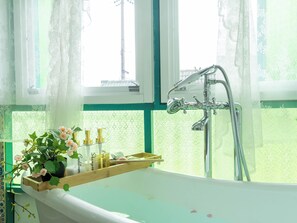 A bathroom with a bathtub, a window with lace curtains, and toiletries on a wooden tray.