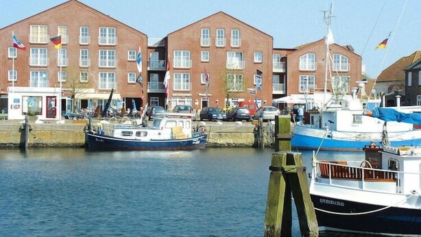 A harbor with boats docked, a brick building with flags, and a wooden post in the foreground. - Ferienwohnung auf Fehmarn in Orther Reede (Orth)