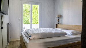 Een slaapkamer met een houten bed, wit beddengoed, een bedlampje en een raam met uitzicht op groen.
