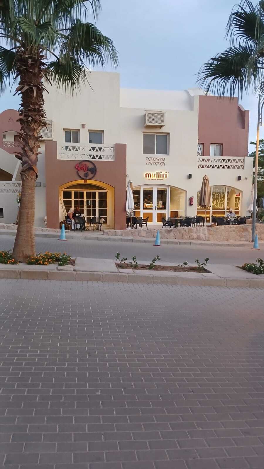 A building with a yellow entrance, outdoor seating, palm trees, and a paved area.