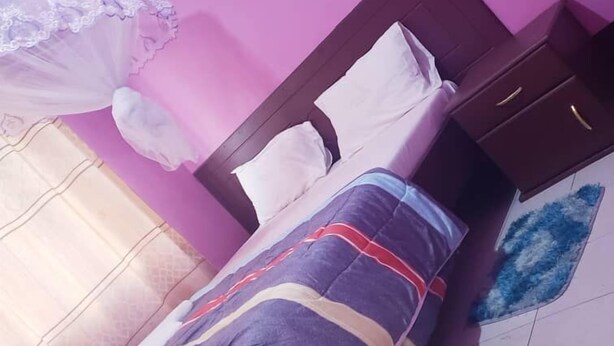 A bedroom with a bed, a nightstand, a dresser, and a window with curtains. - Hotel Delux (Mwanza)