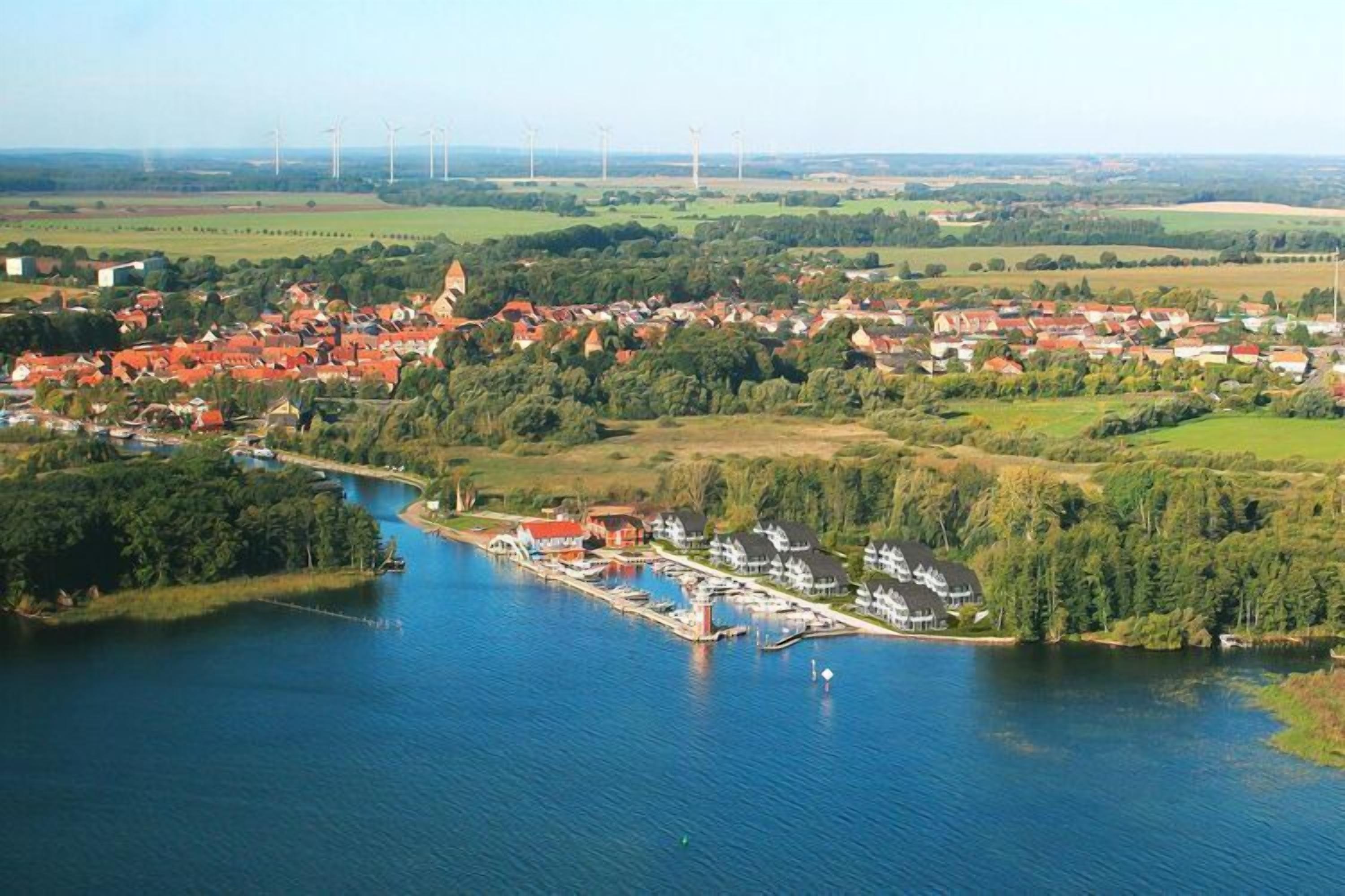 An aerial view of a lakeside village with houses, a marina, and wind turbines in the background.