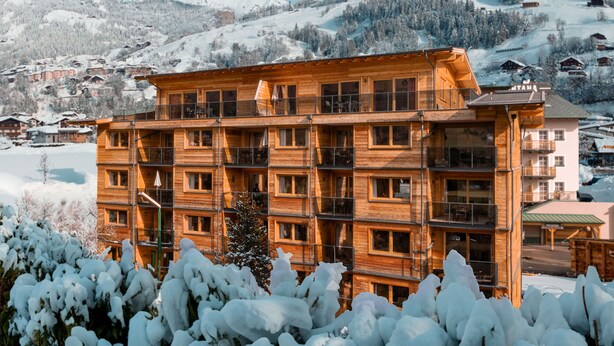 Un immeuble en bois à plusieurs étages, doté de balcons, entouré d’arbres couverts de neige et avec une chaîne de montagnes en arrière-plan.