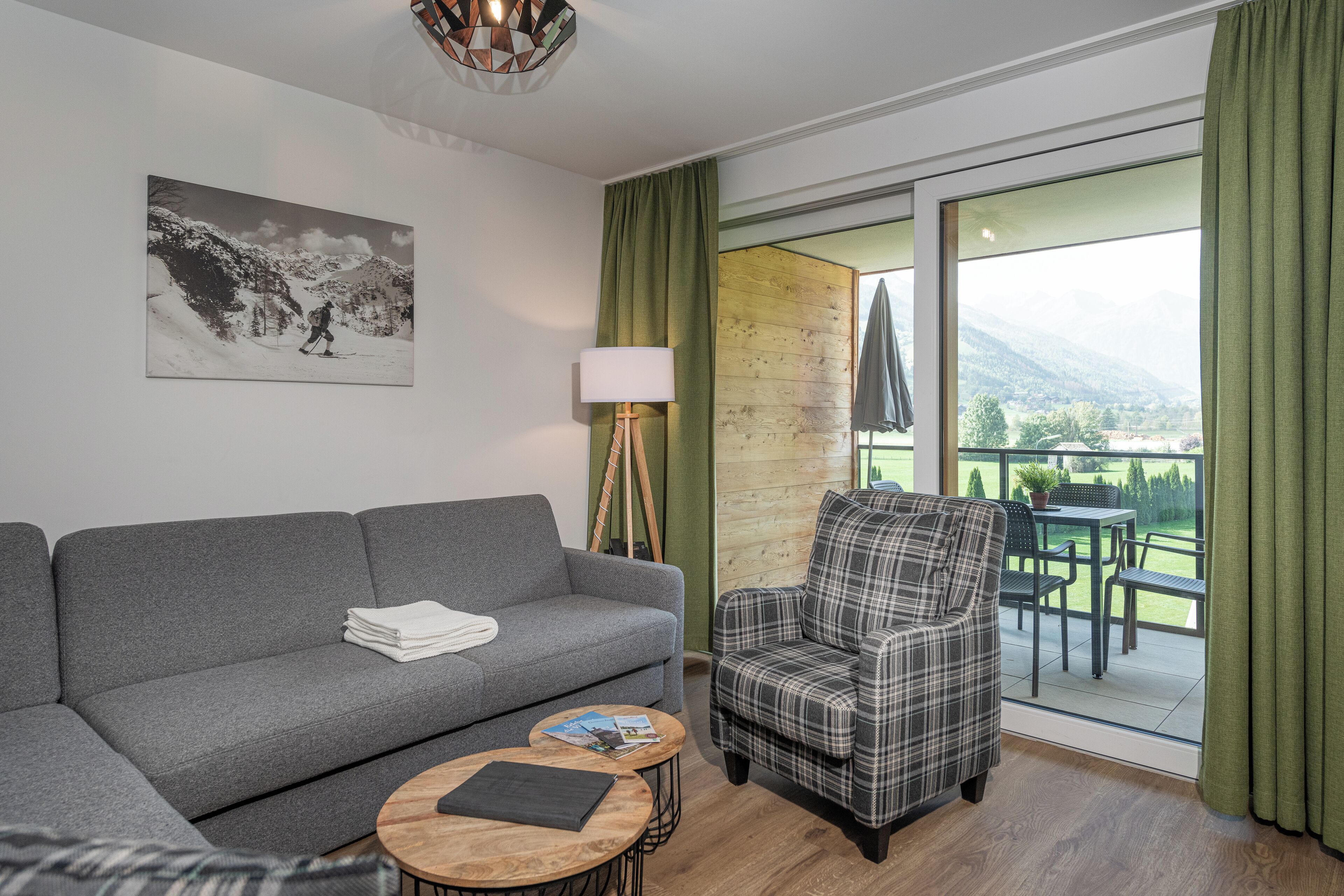 Un salon moderne avec un canapé gris, un fauteuil à carreaux, une petite table ronde et une grande fenêtre donnant sur un paysage verdoyant et des montagnes.