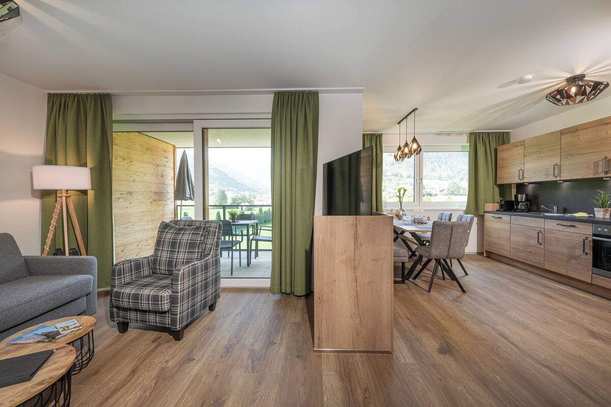 Ein modernes Wohnzimmer mit einer Couch, einem Karoschaukelstuhl, einem Esstisch mit Stühlen, einer Küchenzeile mit Holzschränken und einem großen Fenster mit Vorhängen.