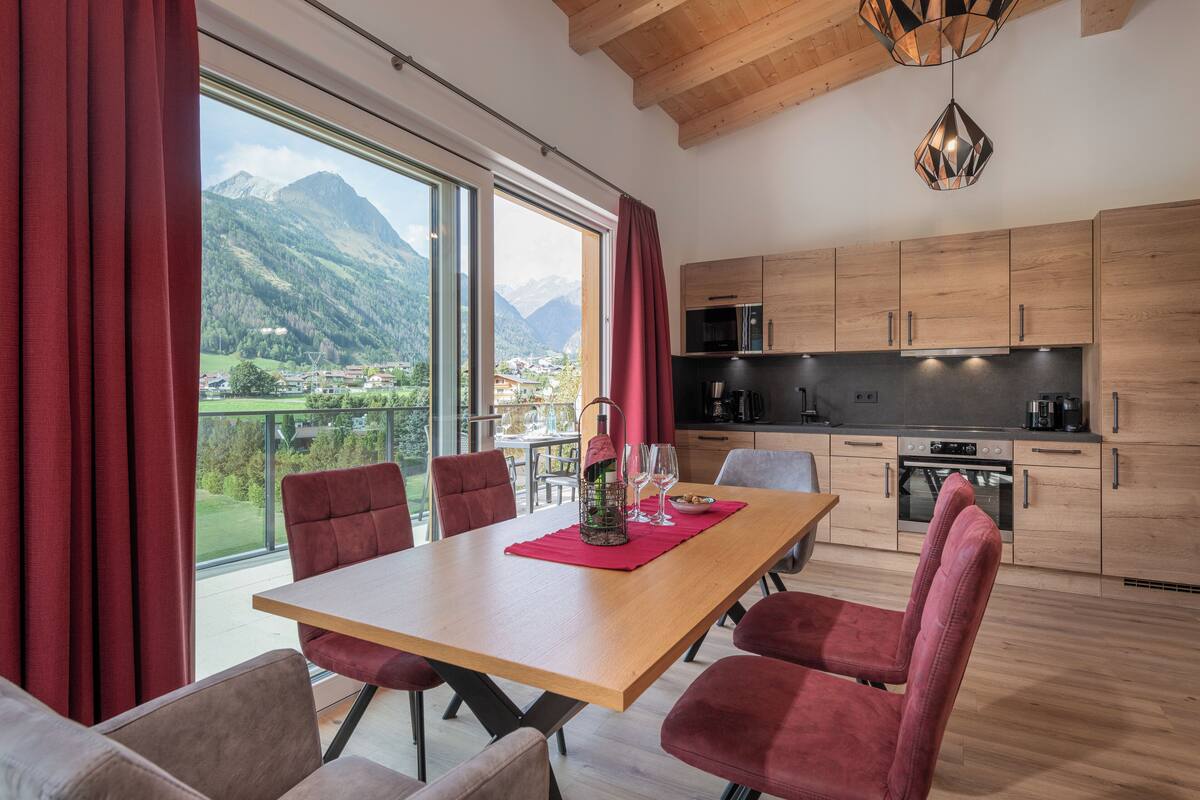 Eine moderne Küche mit Holzausziehtischen und einer großen Kochinsel, ein Essbereich mit roten Stühlen und Blick auf Berge durch große Fenster.