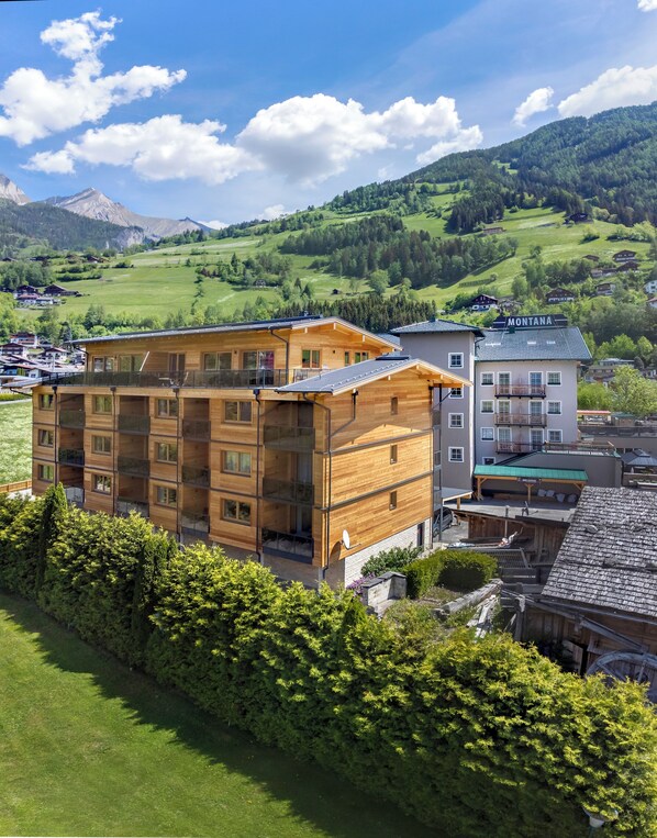 Un edificio de madera de varios pisos con balcones, un letrero que dice "MONTANA" y una vista de colinas y montañas verdes al fondo.