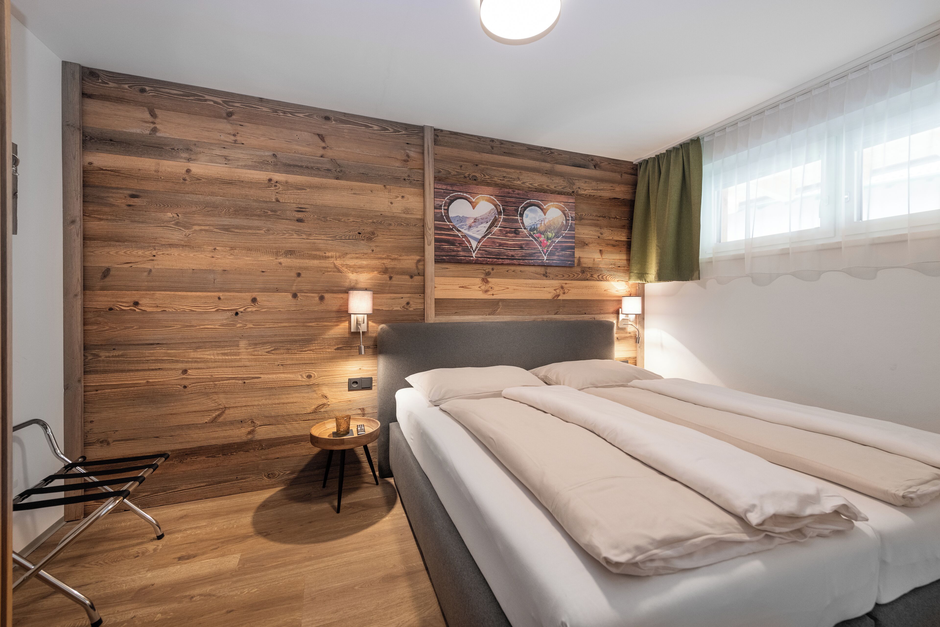 A bedroom with a wooden wall, a bed with white bedding, two wall-mounted lights, and a small table with a lamp.