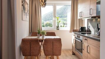 A modern kitchen with wooden cabinets, a dining table with two chairs, and a window with a view of a mountainous landscape.