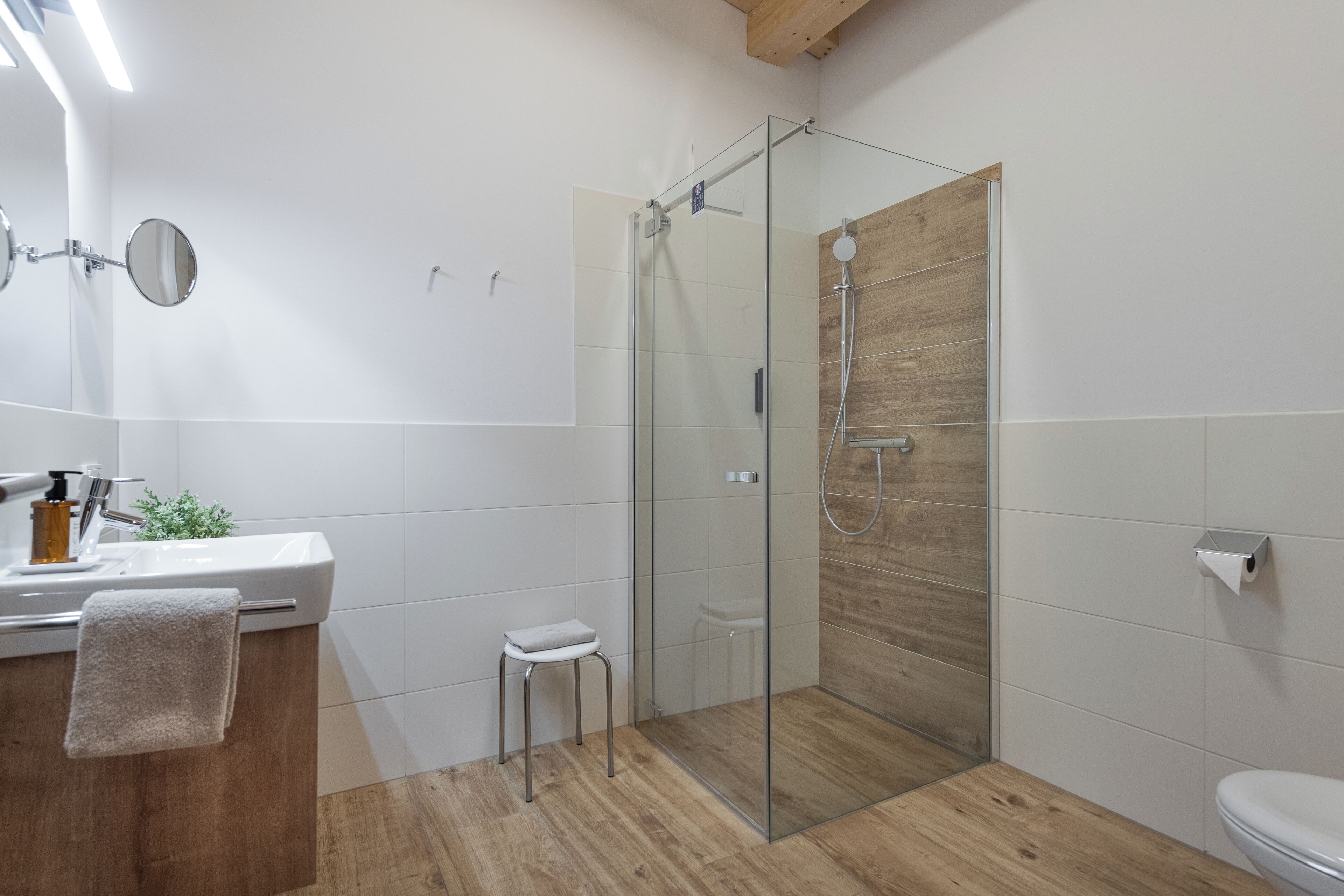 A modern bathroom with a glass shower enclosure, a wooden floor, a white sink with a towel, and a small stool.