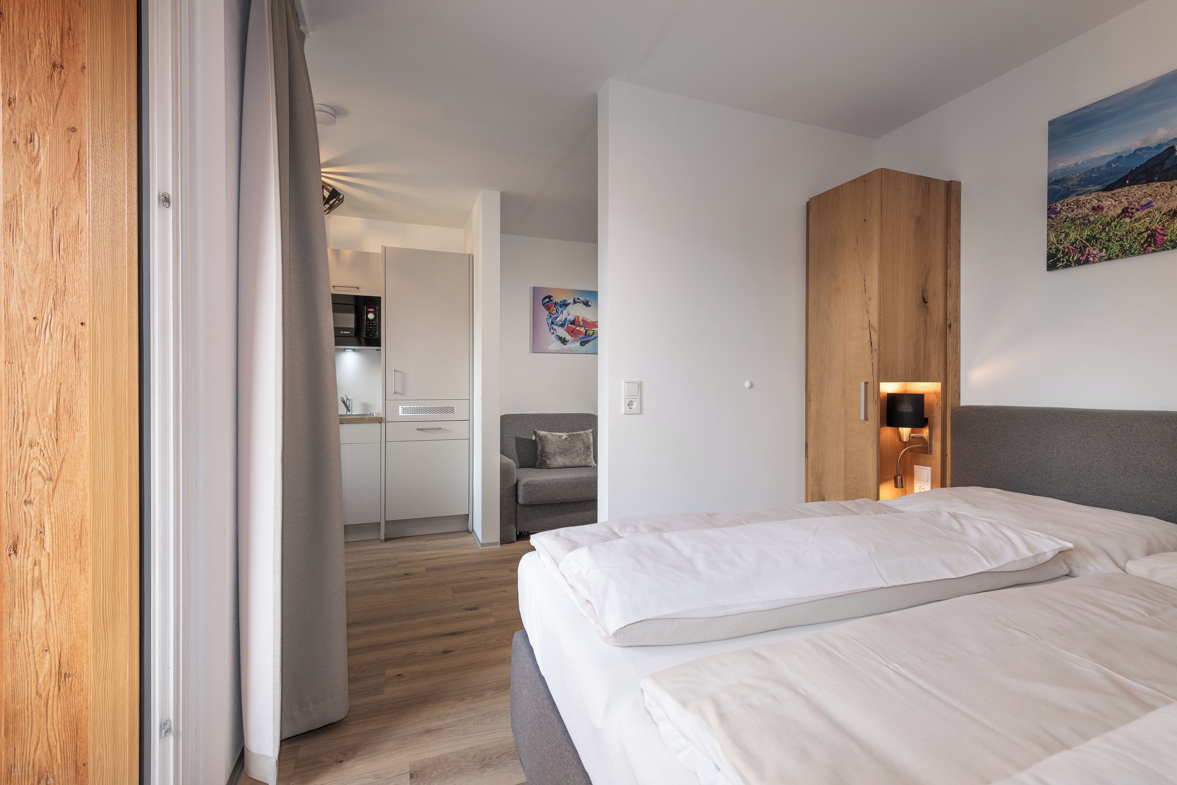 Ett modernt hotellrum med en garderob i trä, en säng med vita sängkläder, ett skrivbord med en stol och en tavla på väggen.