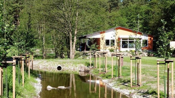 Ein Gebäude am Flussufer mit überdachter Terrasse, einem kleinen Wasserlauf und umgebender Bepflanzung.