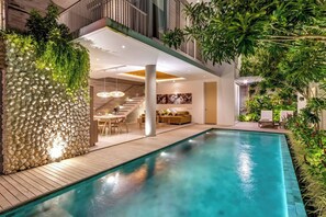A modern indoor pool area with a wooden deck, a stone wall with greenery, and a seating area with a sofa and chairs. - Swan Villas in Seminyak (Seminyak)