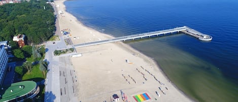 An aerial view of a beach with a long pier extending into the water, a sandy beach area with a rainbow-colored structure, and buildings adjacent to the beach.
