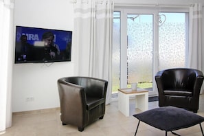 A living room with a flat-screen TV, two leather chairs, a small table, and a view of a garden through a glass door.