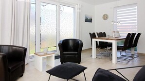 A modern living room with a dining area, featuring a sofa, chairs, a table, and large windows with curtains. - Ferienhaus, Boiensdorf (Boiensdorf)