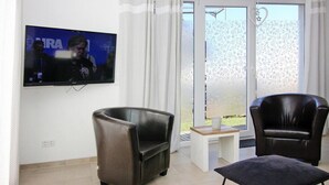 Een woonkamer met een flatscreen televisie, twee leren fauteuils, een kleine tafel en uitzicht op een tuin door een glazen deur.