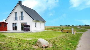 Une maison blanche à deux étages, avec un toit noir, un barbecue abrité et un grand rocher dans le jardin.