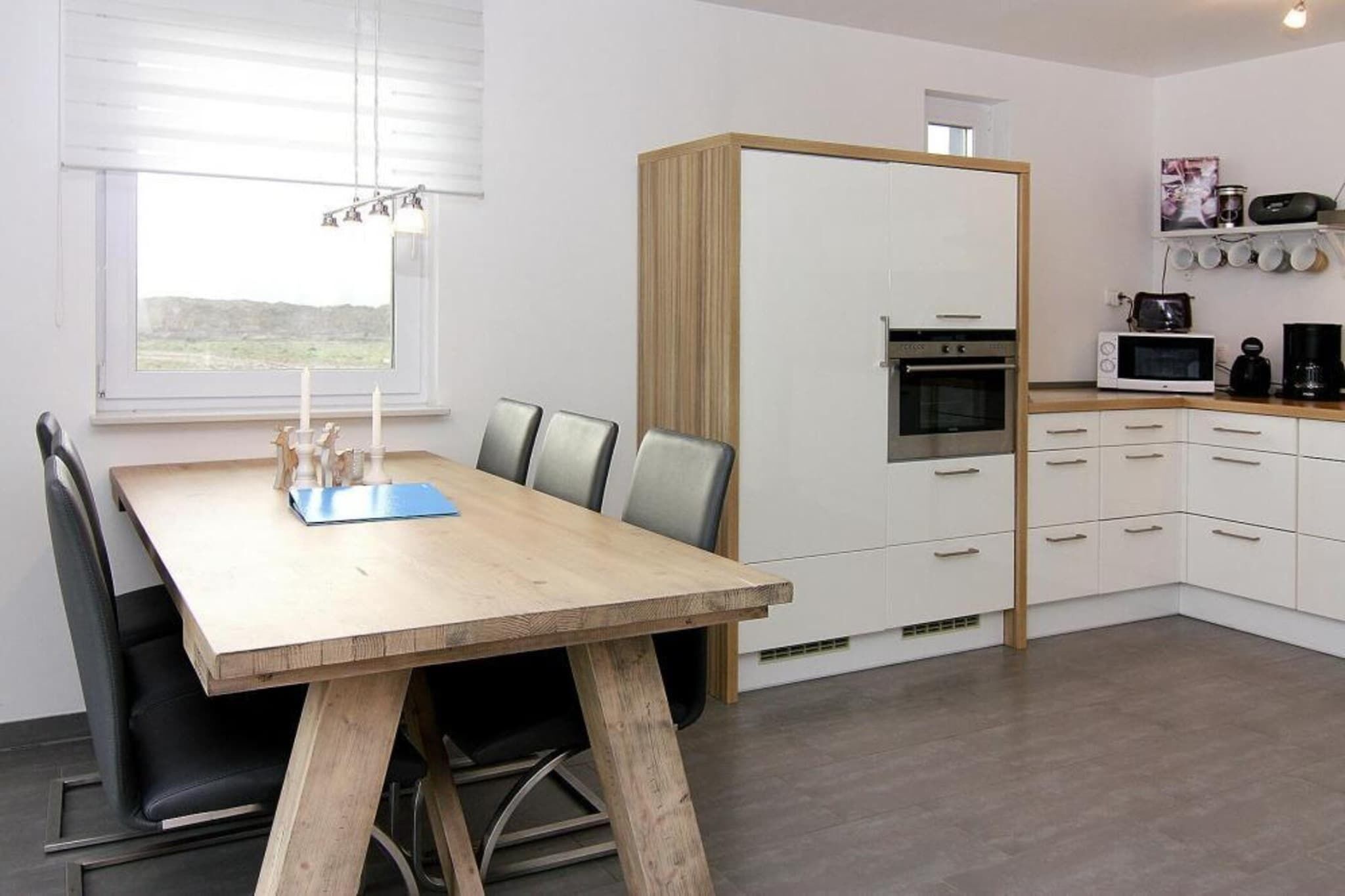 Une cuisine moderne avec des meubles blancs, une table à manger en bois et des chaises, et une vue sur l’extérieur grâce à une grande fenêtre.