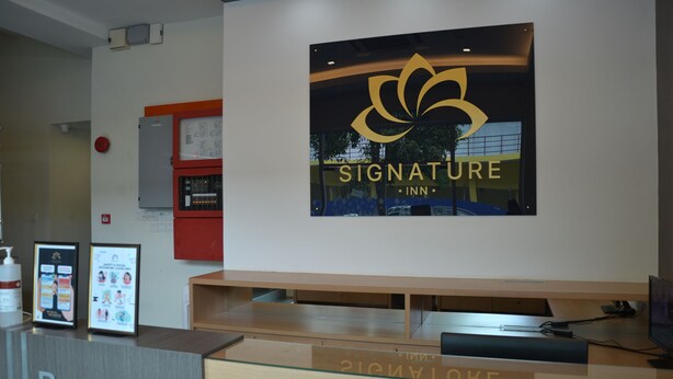 A reception desk with a 'Signature Inn' logo, a reception area, and a wall-mounted fire alarm. - Signature Inn (Kota Kinabalu)