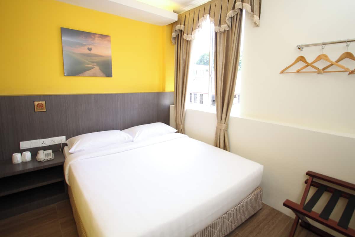 A hotel room with a bed, a bedside table with a phone and cups, a painting on the wall, and a window with curtains.