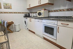 A kitchen with a washing machine, microwave, oven, and a wooden chair. - 15A06 Apartamento centrico en Llanes (Llanes)