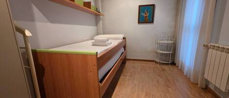 A small room with a wooden bunk bed, a shelf with decorative items, a framed picture on the wall, and a radiator.