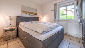 Ein Schlafzimmer mit Bett, Nachttisch, Lampe, Fenster und einem Kunstwerk an der Wand.