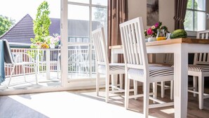A dining area with white chairs and a table, a glass door leading outside, and a view of a house and trees. - Wohnung in Zingst nahe Ostseestrand (Zingst)