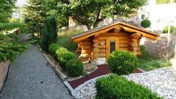 Eine Holzhütte mit Holztür und kleiner Veranda, umgeben von einem Kiesweg und geschnittenen Büschen.