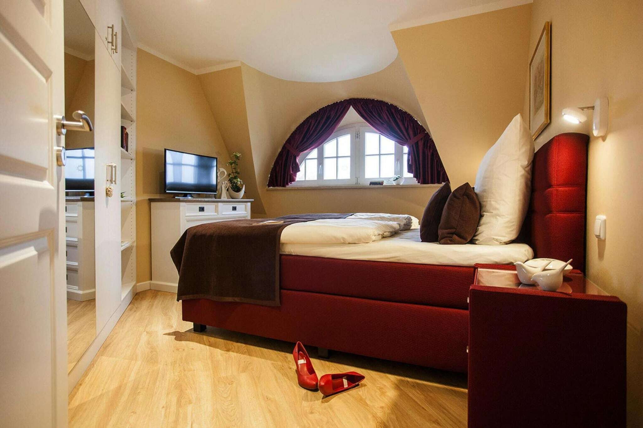 Une chambre avec un lit rouge, un téléviseur posé sur un meuble, une fenêtre avec des rideaux et un plancher en bois.