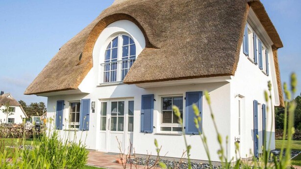 Une maison avec un toit de chaume et des murs blancs, dotée de nombreuses fenêtres ornées de volets bleus.