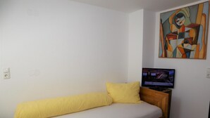 Ein Einzelbett mit gelber Bettwäsche, ein Fernseher auf einem hölzernen Standfuß und ein farbenfrohes, abstraktes Gemälde an der Wand.
