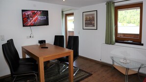 Ein Hotelzimmer mit einem Holztisch, schwarzen Stühlen, einem Glastisch, einem an der Wand befestigten Fernseher, einem gerahmten Bild, einem Fenster mit Vorhängen und einem Heizkörper.
