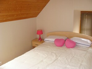 Quarto com teto de madeira, uma mesa de cabeceira individual com abajur vermelho, cama com roupa de cama branca e dois travesseiros em formato de coração, e uma janela com persianas.