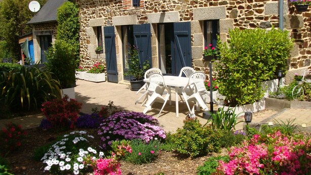 A stone building with a garden in front. There are white chairs and a table in the garden.