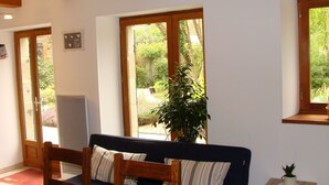 A cozy living room with a dark blue sofa, wooden chairs, a small table with a plant, and large windows offering a view of a garden.