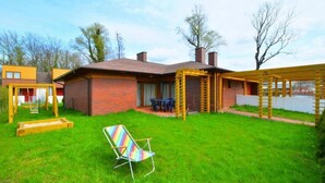 Un patio trasero con una casa de ladrillo, una estructura de juego de madera, una silla de colores y una pérgola de madera.